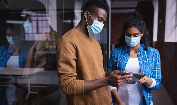 Diverse male and female colleagues at work discussing and looking at tablet wearing face masks. independent creative design business during covid 19 coronavirus pandemic.