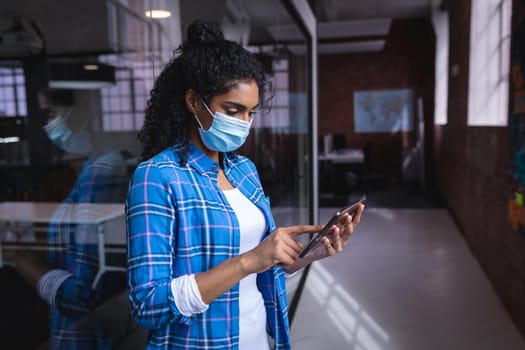 Mixed race businesswoman wearing face mask standing in corridor looking at tablet. independent creative design business during covid 19 coronavirus pandemic.