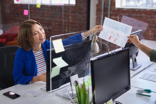 Mixed race female in front of computers separated by sneeze shield giving document to colleague. independent creative design business during covid 19 coronavirus pandemic.