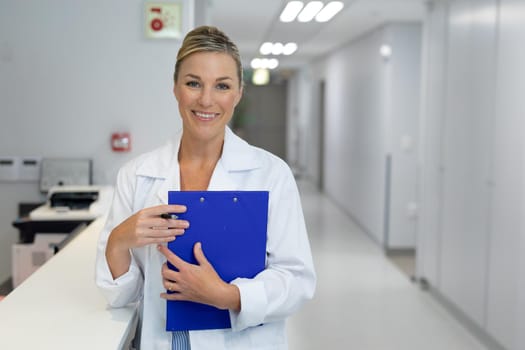 Portrait of happy caucasian female doctor standing in hospital corridor holding medical document. medicine, health and healthcare services.