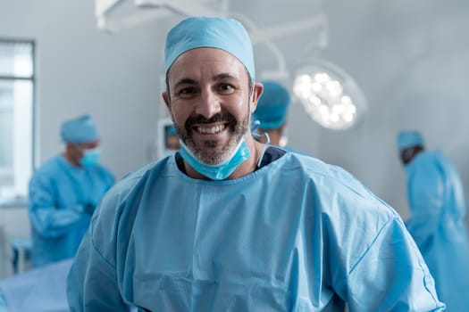 Smiling caucasian male surgeon with face mask and protective clothing in operating theatre. medicine, health and healthcare services during covid 19 coronavirus pandemic.