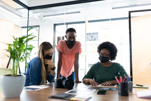 Diverse group of business colleagues wearing masks brainstorming looking at photos in meeting room. independent creative business during covid 19 coronavirus pandemic.
