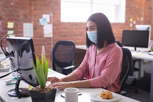 Mixed race businesswoman sitting in office in front of computer wearing face mask. independent creative design business during covid 19 coronavirus pandemic.