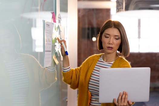 Asian businesswoman standing in front of whiteboard and writing holding laptop having video call. independent creative design business.