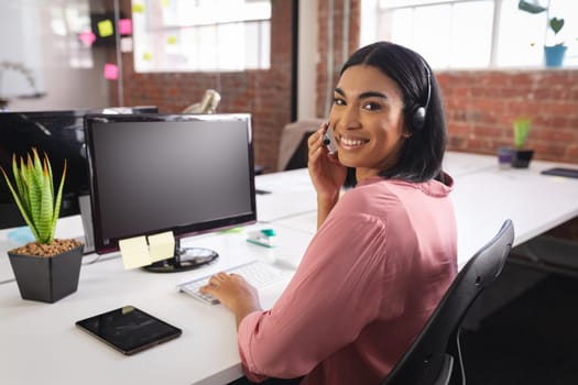 Happy mixed race businesswoman having video call sitting in front of computer using headphones. independent creative design business.