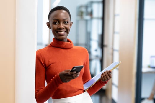 Happy african american businesswoman standing in corridor holding smartphone and documents. business in a modern office.
