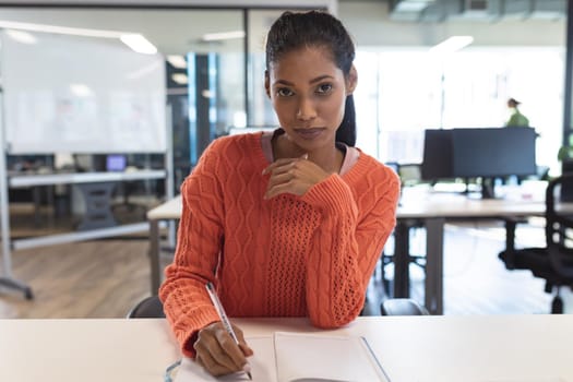 Portrait of mixed race female creative worker sitting at desk looking at camera. modern office of a creative design business.