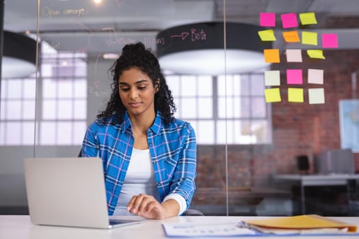 Mixed race businesswoman sitting in front of laptop with sticky memos on wall in background. independent creative design business.