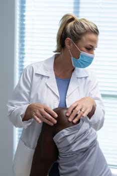 Caucasian female physiotherapist wearing face mask palpating knee of african american male patient. medicine, health and healthcare services during coronavirus covid 19 pandemic.