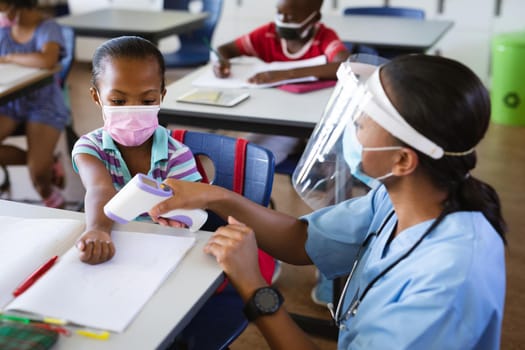 Female health worker wearing face shield measuring temperature of a girl at elementary school. education back to school health safety during covid19 coronavirus pandemic.