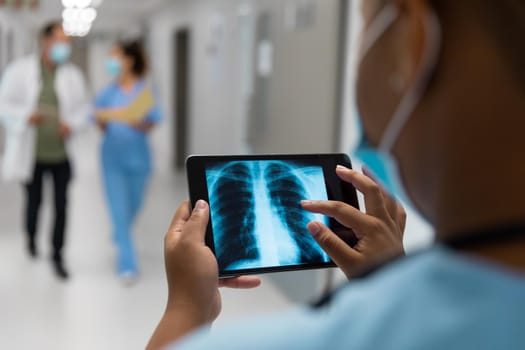 Mixed race doctor in corridor wearing face mask looking at lung x-ray on tablet. medicine, health and healthcare services during coronavirus covid 19 pandemic.