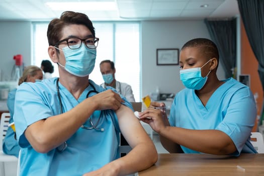 African american female doctor giving covid vaccination to male colleague, both in face masks. medicine, health and healthcare services during covid 19 coronavirus pandemic.