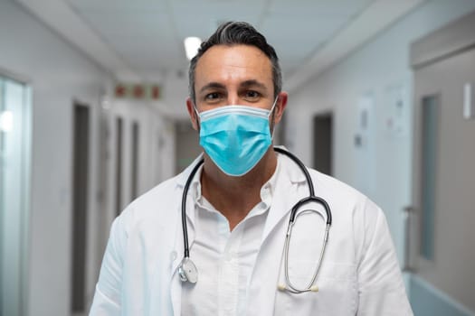 Portrait of mixed race male doctor wearing face mask standing in hospital corridor. medicine, health and healthcare services during coronavirus covid 19 pandemic.
