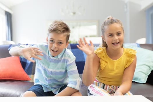 Caucasian brother and sister waving and smiling during video chat at home. domestic lifestyle and leisure family time concept.