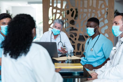 Caucasian female senior doctor and diverse hospital colleagues wearing face masks sitting at meeting. medicine, health and healthcare services during covid 19 coronavirus pandemic.