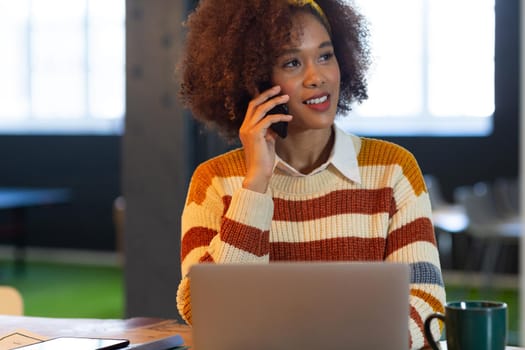 Mixed race businesswoman sitting at conference table with laptop and talking on smartphone. independent creative design business.