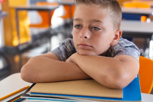 Tired looking caucasian schoolboy sitting at desk in classroom leaning on a pile of schoolbooks. childhood and education at elementary school.