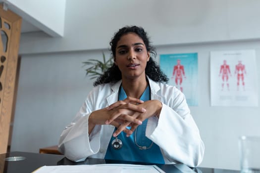 Mixed race female doctor at desk talking and gesturing during video call consultation. telemedicine, online healthcare services during quarantine lockdown