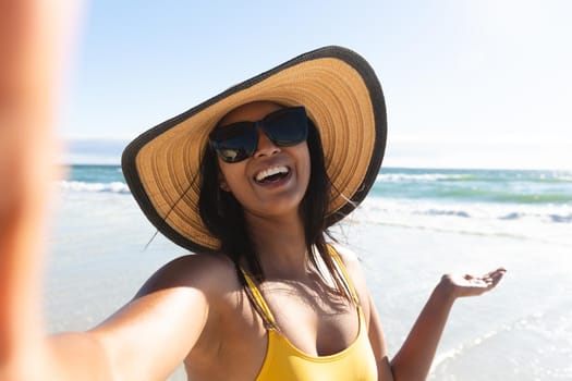 Smiling mixed race woman wearing sunhat and sunglasses taking selfie on beach. summer vacation beach holiday break.