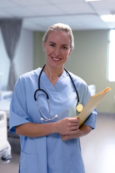 Portrait of smiling caucasian female doctor in hospital wearing scrubs and stethoscope holding files. medical professional at work and health services.