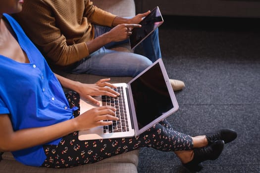Diverse male and female colleagues working sititng on sofa in office using computer and tablet. independent creative design business.