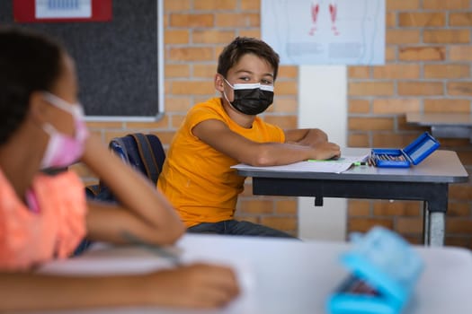 Caucasian boy wearing face mask sitting on his desk in the class at elementary school. back to school and education concept