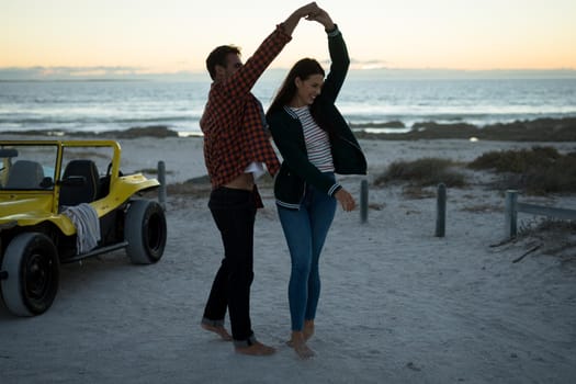 Happy caucasian couple next to beach buggy by the sea dancing. beach break on summer holiday road trip