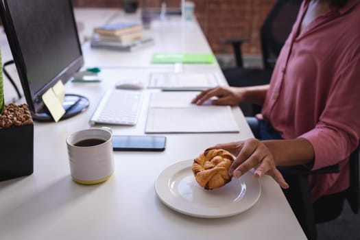 Mixed race businesswoman sitting in office in front of computer and having snack. independent creative design business.