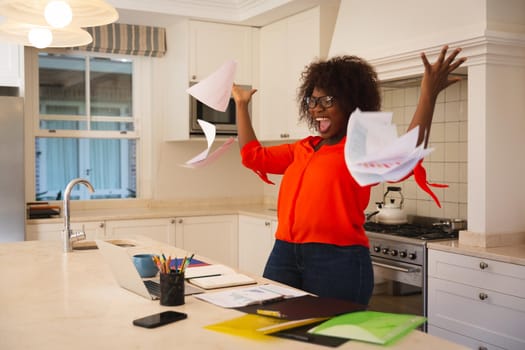 Happy african american woman working in kitchen with laptop, celebrating success, throwing papers. flexible working from home.