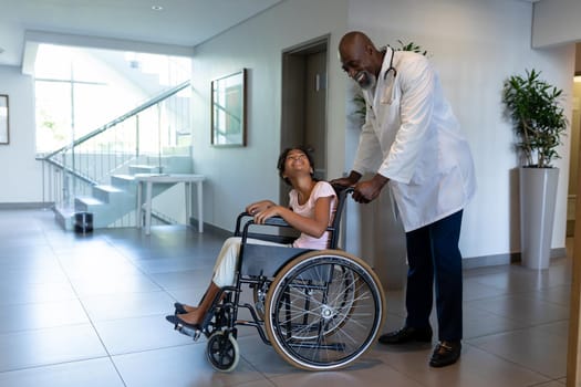 Smiling african american male doctor pushing mixed race girl in wheelchair through hospital corridor. medicine, health and healthcare services.