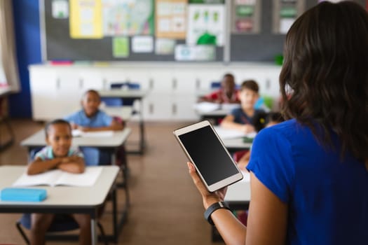 Rear view of female teacher holding digital tablet teaching students in the class at school. school and education concept