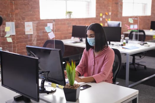 Mixed race businesswoman sitting in office in front of computer wearing face mask. independent creative design business during covid 19 coronavirus pandemic.