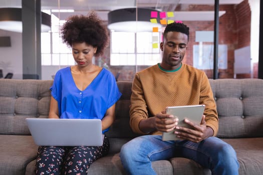 Diverse male and female colleagues working sititng on sofa in office using computer and tablet. independent creative design business.