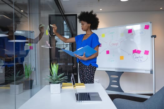 Mixed race businesswoman sticking memo notes on glass wall with whiteboard in background. independent creative design business.