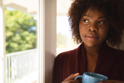 Thoughtful african american woman looking out of sunny window holding cup of coffee. spending free time at home.