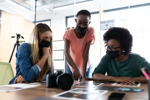 Diverse group of business colleagues wearing masks brainstorming looking at photos in meeting room. independent creative business during covid 19 coronavirus pandemic.