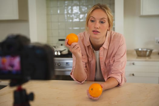 Caucasian woman in kitchen holding oranges and talking to camera, making cooking vlog. technology and communication, cookery vlogger at home.