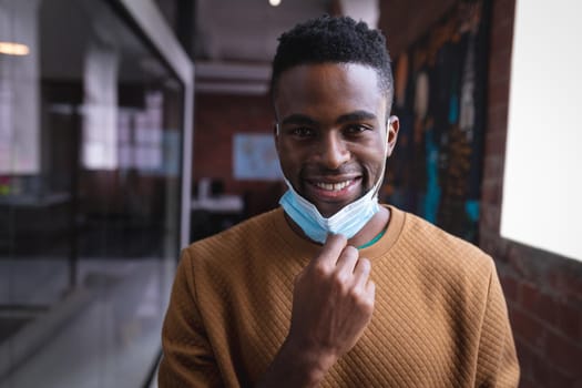 Portrait of happy african american businessman taking off face mask standing in corridor in office. independent creative design business during covid 19 coronavirus pandemic.