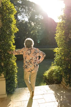 Senior caucasian woman leaning on column in sunny garden. luxury retirement lifestyle, spending time alone at home.