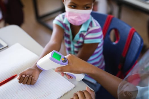 Mid section of female health worker measuring temperature of a girl at elementary school. education back to school health safety during covid19 coronavirus pandemic.
