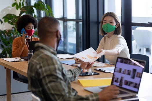 Diverse group of business people working in creative office. group of people wearing face masks and giving documents. social distancing protection hygiene in workplace during covid 19 pandemic.