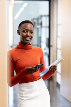 Happy african american businesswoman standing in corridor holding smartphone and documents. business in a modern office.