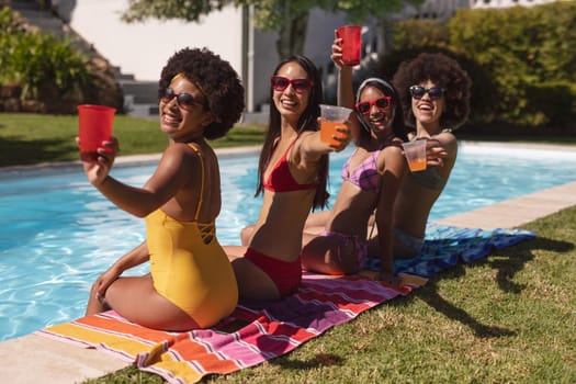 Diverse group of female friends drinking drinks sitting at the poolside. Hanging out and relaxing outdoors in summer.