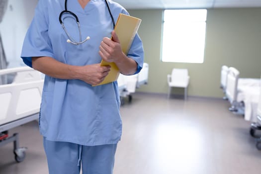 Midsection of caucasian female doctor in hospital wearing scrubs and stethoscope holding files. medical professional at work and health services.