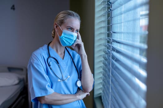 Worried caucasian female doctor in hospital wearing face mask and scrubs looking out of window. medical professional at work and health services during coronavirus covid 19 pandemic.
