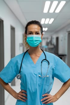 Portrait of mixed race female doctor wearing face mask standing in hospital corridor. medicine, health and healthcare services during coronavirus covid 19 pandemic.