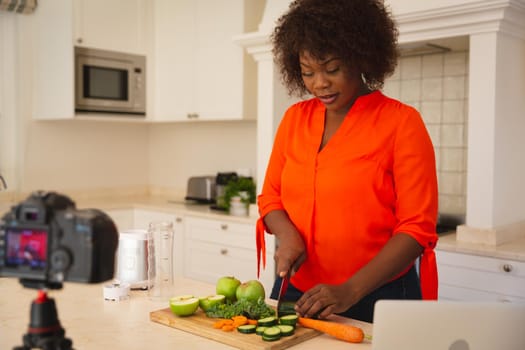 Happy african american woman standing in kitchen preparing food, making vlog using laptop and camera. online cookery vlogger at home.