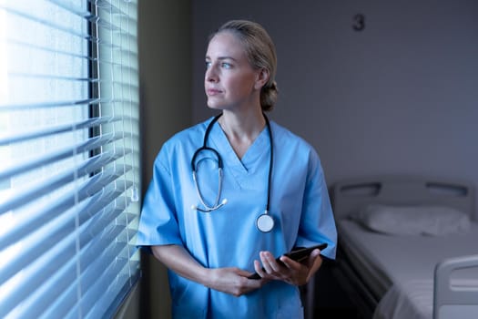 Pensive caucasian female doctor in hospital wearing scrubs and stethoscope, looking out of window. medical professional at work and health services.