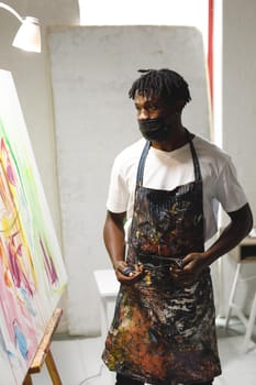 African american male painter wearing face mask painting on canvas in art studio. creation and inspiration at an artists painting studio during coronavirus covid 19 pandemic.