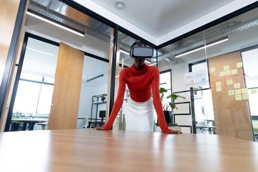 African american businesswoman leaning on desk and using vr headset in meeting room. business in a modern office.